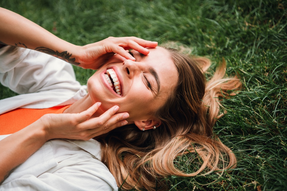 A cheerful young woman lies in green grass on a sunny day, smiling and enjoying a moment of leisure. This lifestyle image captures happiness, fashion, and outdoor park charm.