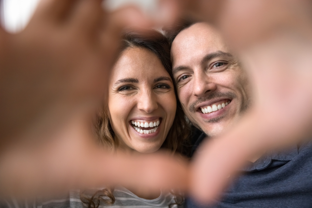 Beautiful Hispanic wife and husband look at camera through fingers showing symbol of love, close up faces view. 