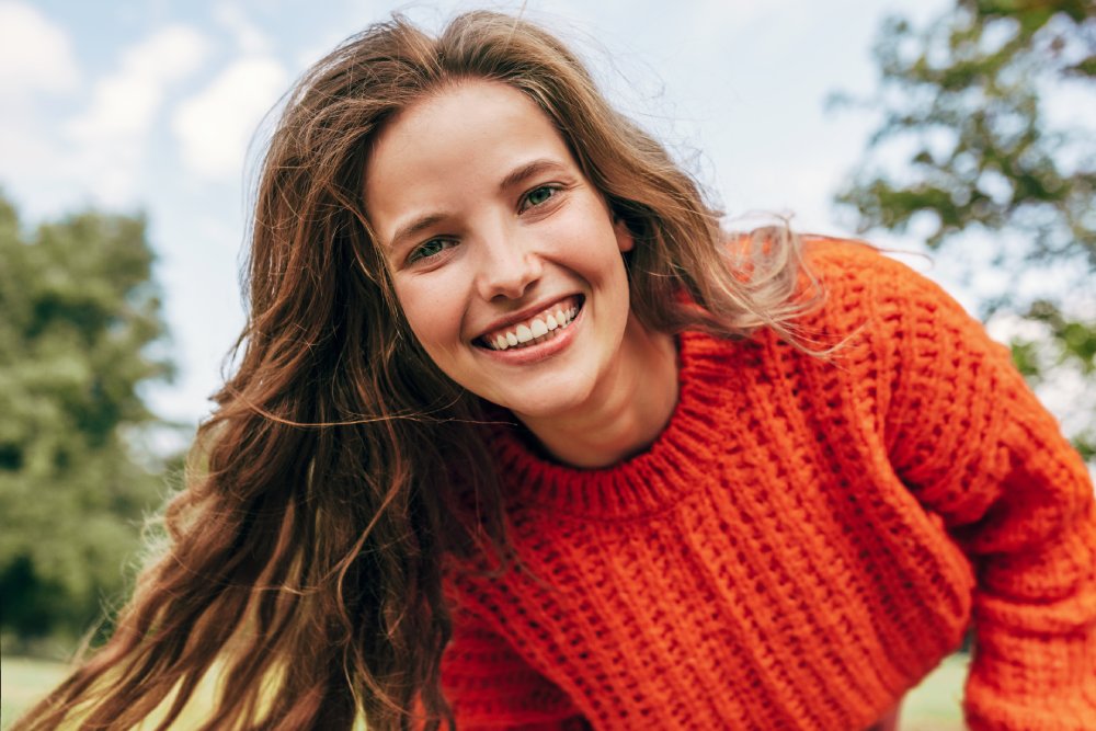 Women’s Health Logo Representing Compassionate Care – obgyn miami Candid portrait of a cheerful young woman wearing an orange sweater spending time in the park. The pretty female has a joyful expression, resting outdoors on nature background.
