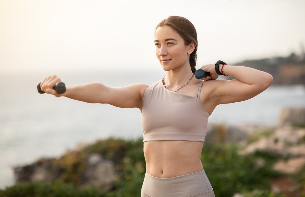 Glad confident strong millennial caucasian woman athlete in sportswear doing arm exercises with dumbbells, enjoy workout, body care alone at sea beach. Sports, fitness outdoor
