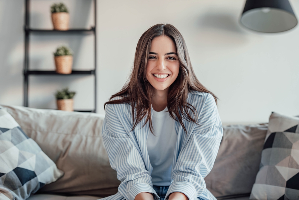 Young attractive cheerful lady sitting on sofa, relaxing looking at the camera at home. Beautiful brunette woman on her 30s chatting on video call online